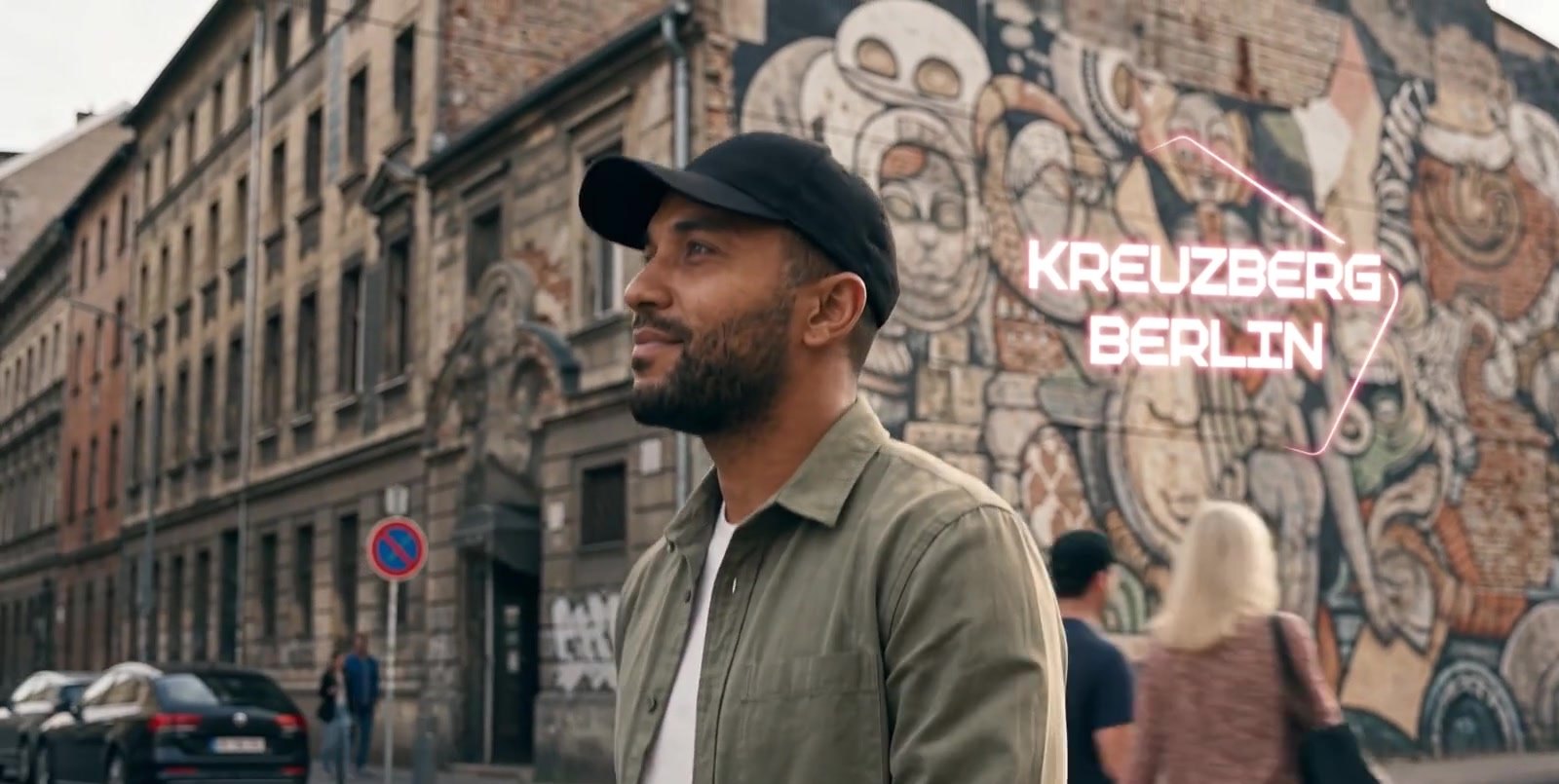Film frame: the Builder in front of the Kreuzberg mural, a neon outline reading Kreuzberg Berlin